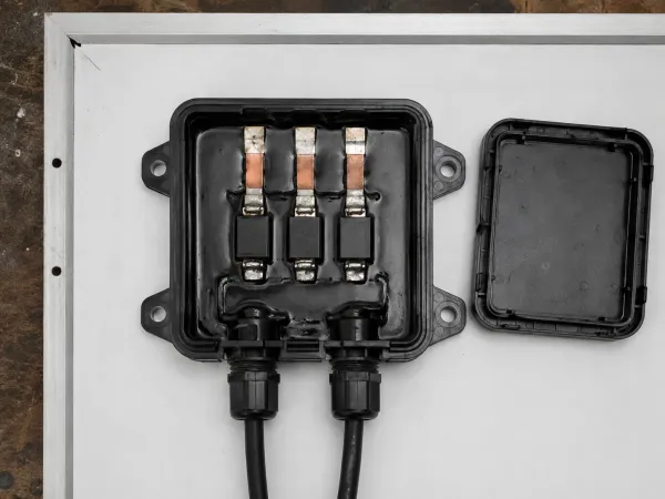 Illustration generated for Solar Power Magazine, 1 Sep 2026. Not a photograph. No real installation, product or document depicted. Illustration: a photovoltaic junction box on the white backsheet of a module, lid removed and set beside it, showing three ribbon tabs entering the potting compound and three bypass diodes.